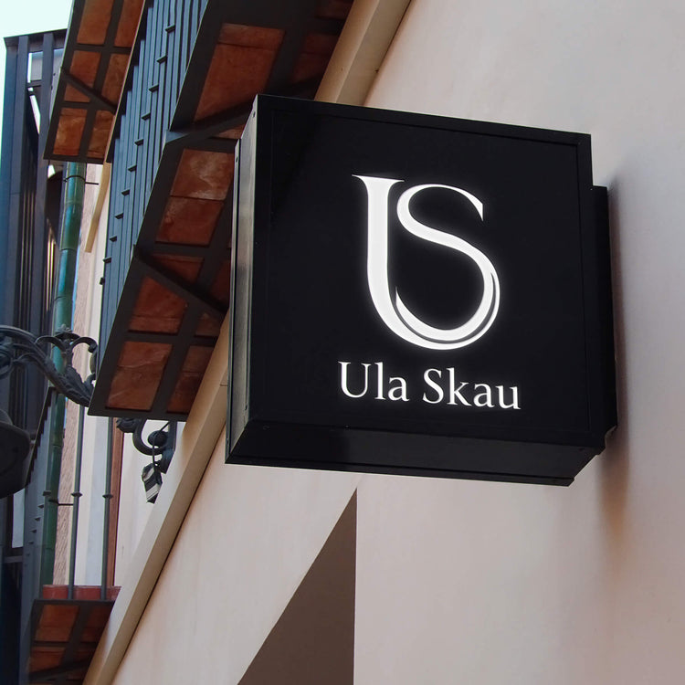 Illuminated sign featuring the 'US' monogram and the name 'Ula Skau' on a black background, displayed on a building exterior.